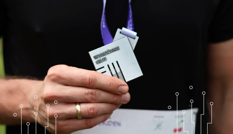 Uczestnik drużyny XEMI Team trzyma medal z tekstem "w drużynie siła" po biegach w sztafecie charytatywnej Plum Ekiden 2025. Na zdjęciu widać tylko kawałek torsu uczestnika w czarnej koszulce i medal, który znajduje się w centralnym miejscu zdjęcia.