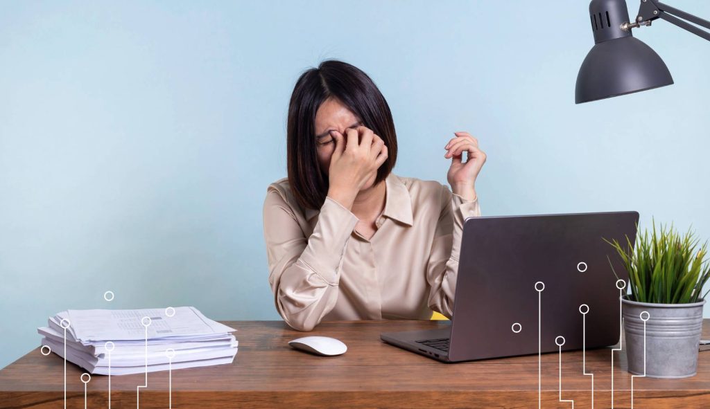 Zestresowana pracowniczka siedzi przy biurku z laptopem i dokumentami, pokazując niepokój związany z nadchodzącą zmianą lub towarzyszący wdrożeniu systemu informatycznego.